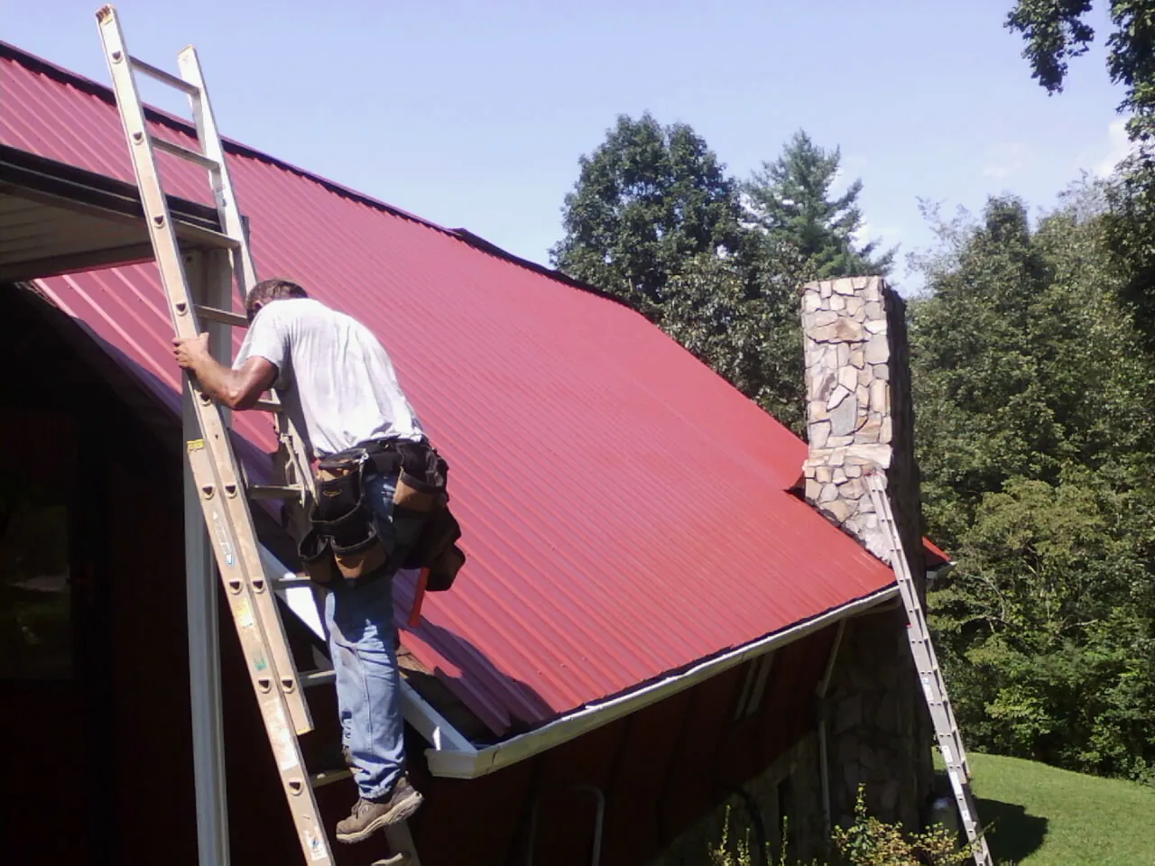 Roofer on a ladder beside a red metal roof and stone chimney — Soffit & Fascia Repair by Roofing Brothers in Comfort